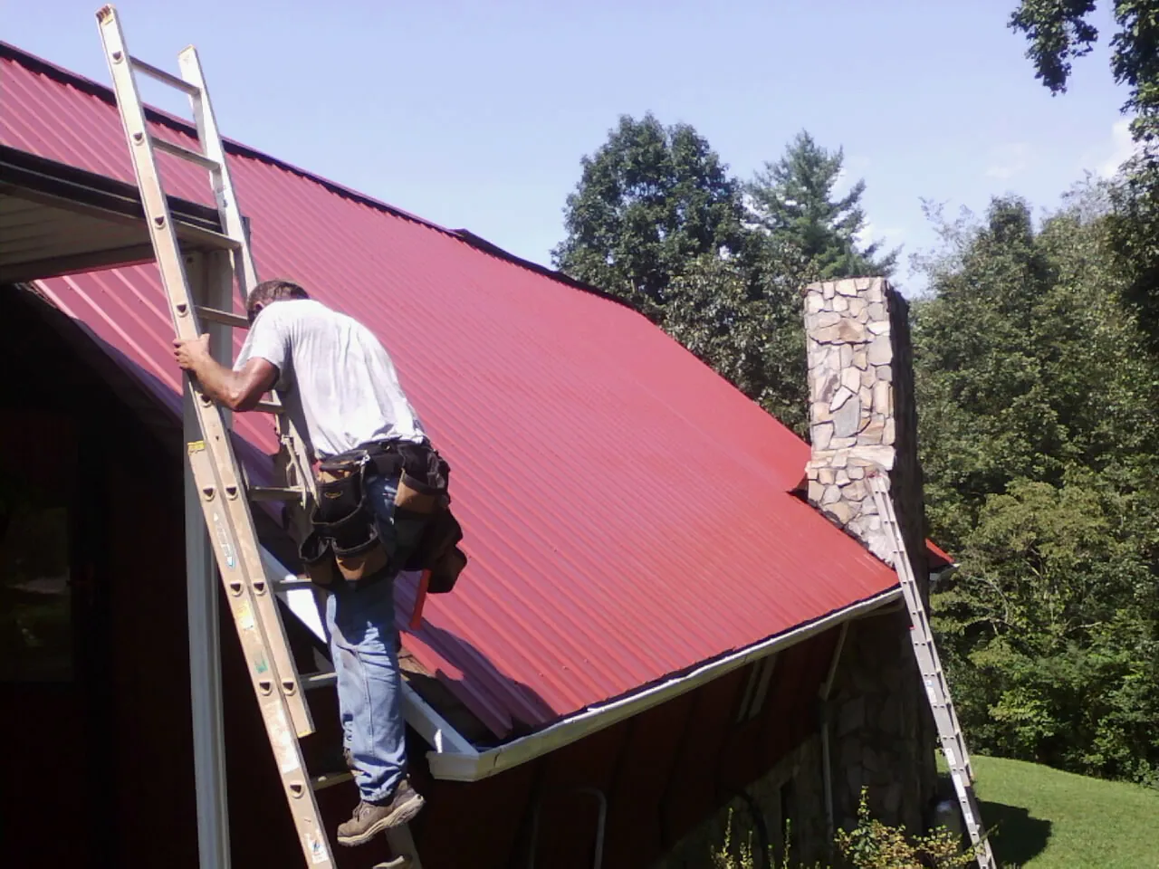 Roofer on a ladder beside a red metal roof and stone chimney — Soffit & Fascia Repair by Roofing Brothers in Dinuba